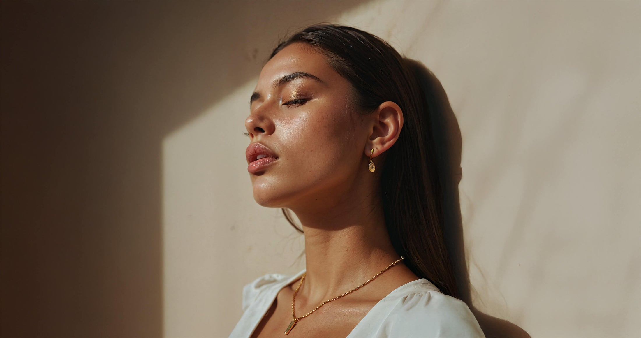 Model with long brown hair and sunkissed skin leans against a wall, bathed in sunlight.